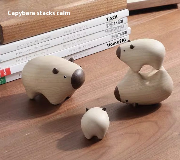 Three light natural wood animal-shaped stacking toys on a wooden tabletop, with two larger pig figures and one smaller pig figure. The text 'Capybara stacks calm' appears in the upper left.