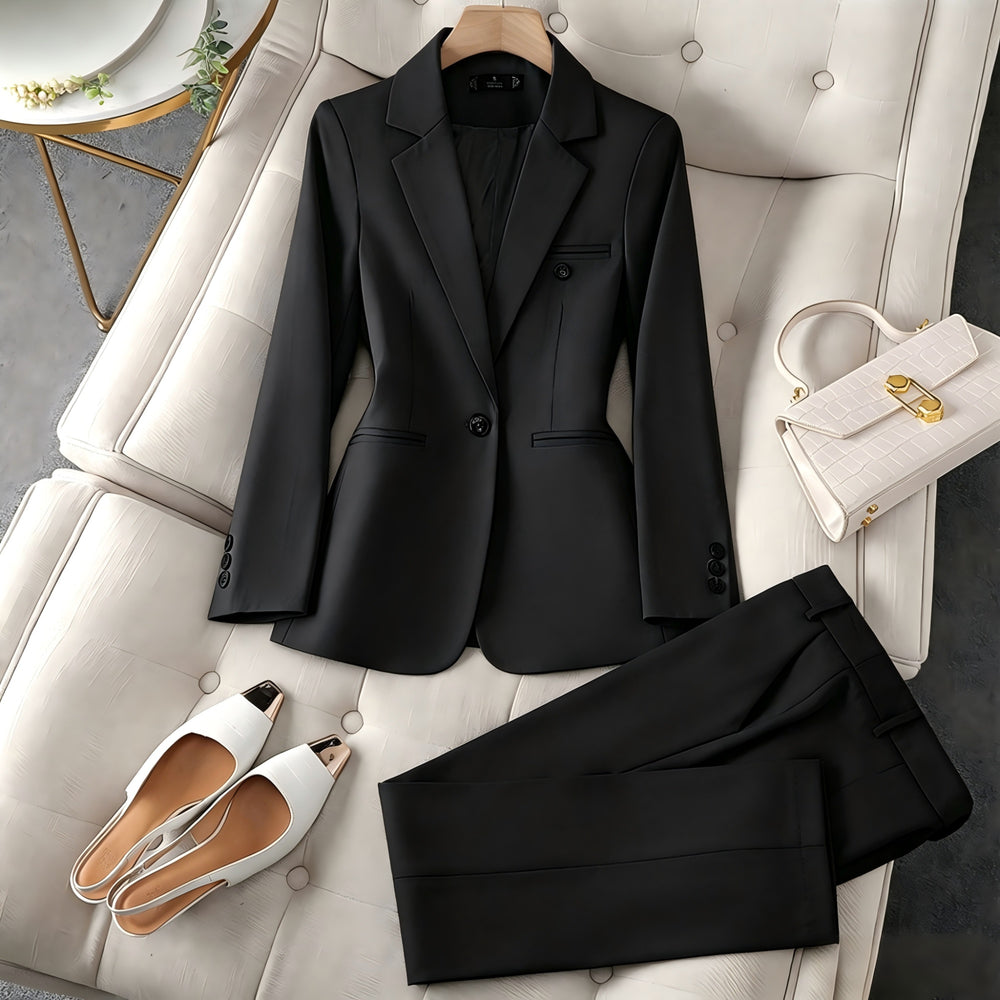 A black tailored blazer and matching trousers are arranged on a cream tufted sofa, styled with white pointed flats, a white handbag, and a small decorative plant in the corner.