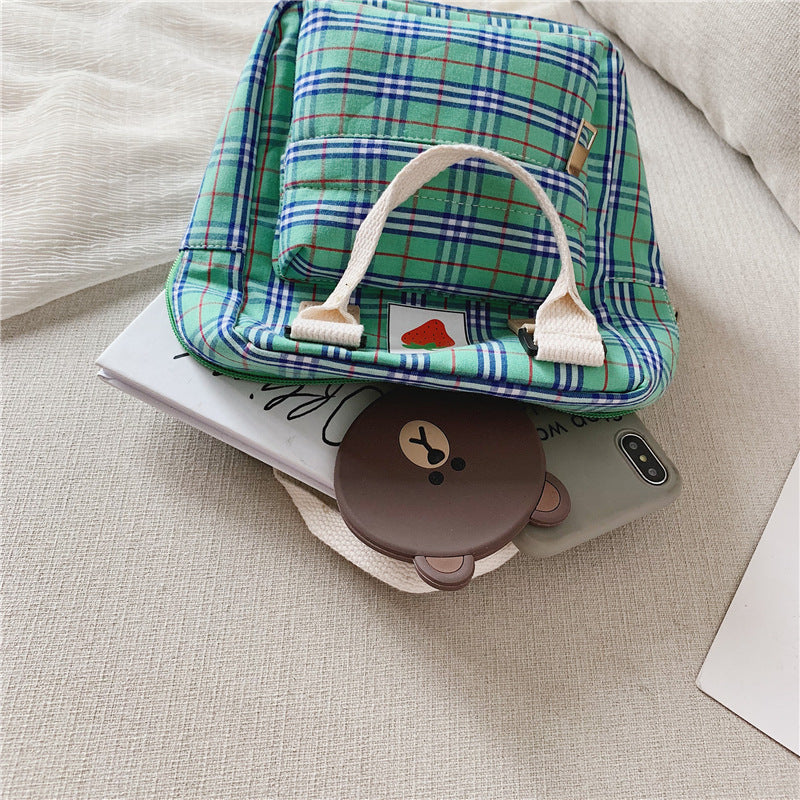 Green plaid bag with a white handle on top of a phone and small round brown object on a beige surface.