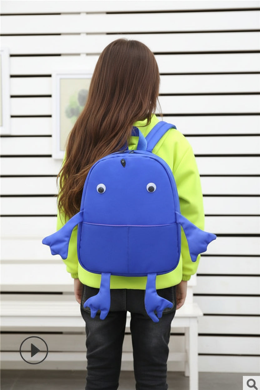 Child wearing a blue backpack shaped like a cartoon character against a white wall.