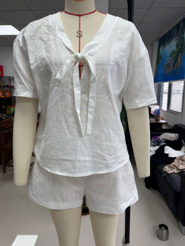 A white short-sleeve linen-like top and matching shorts set displayed on a mannequin torso in an indoor room. The top has a V-neck with a front tie detail, and the mannequin is also wearing a red cord necklace with an S pendant.
