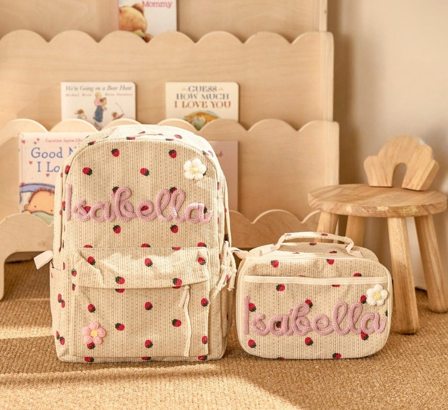 A beige backpack and matching lunch bag set with red polka dots and pink embroidered 'Isabella' text, styled on a woven rug in front of a wooden Kids Corduroy Backpack & Lunch Bag Set – Handmade Chunky Yarn, 7 Colour Combos.