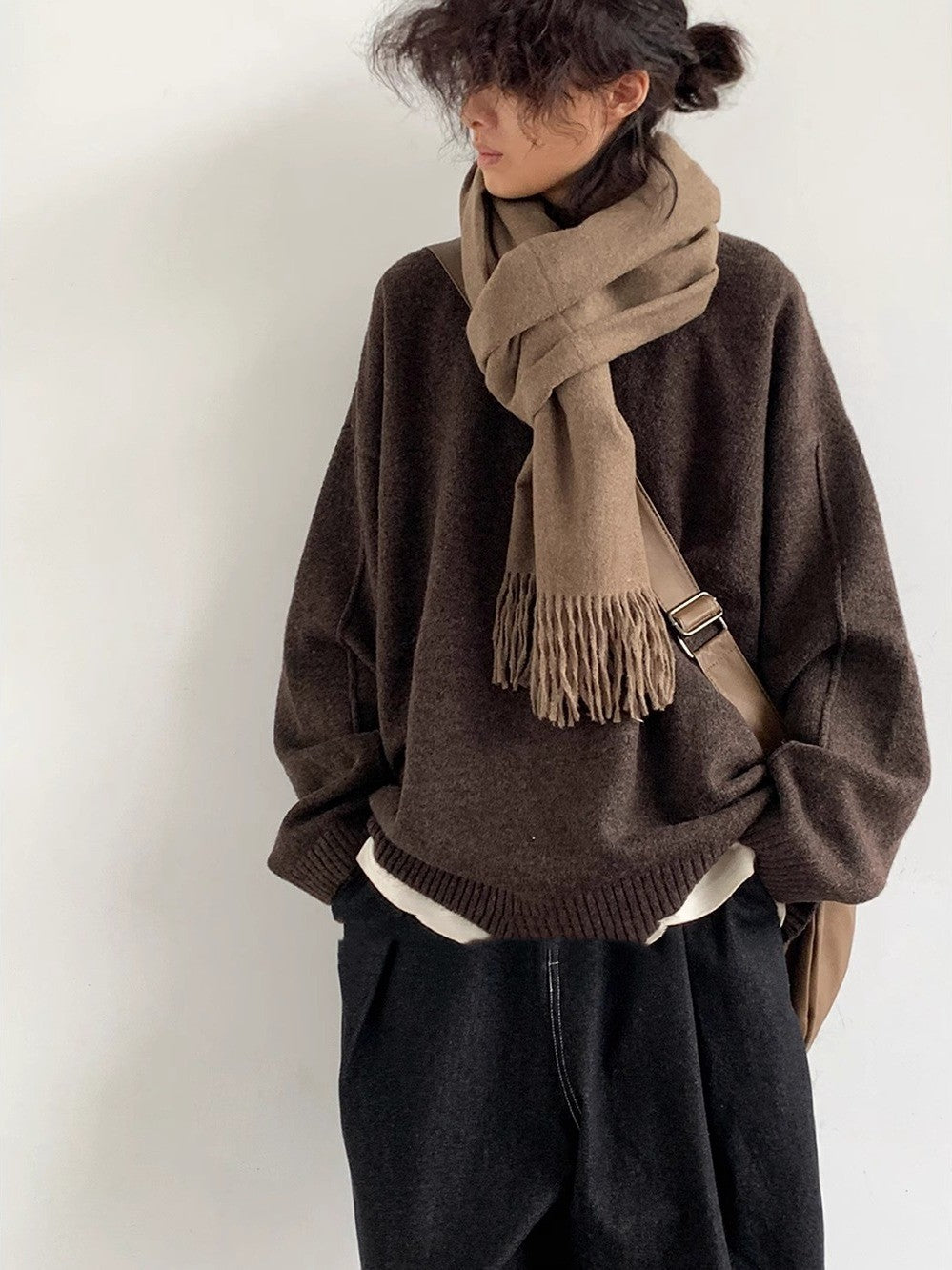 Person wearing a brown sweater and scarf with a beige bag against a white background