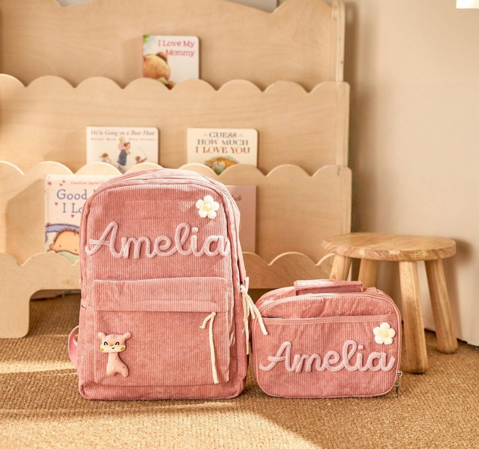 A pink corduroy backpack and matching zip pouch set with the name 'Amelia' embroidered on the front, styled on a beige rug in front of a wooden bookshelf.