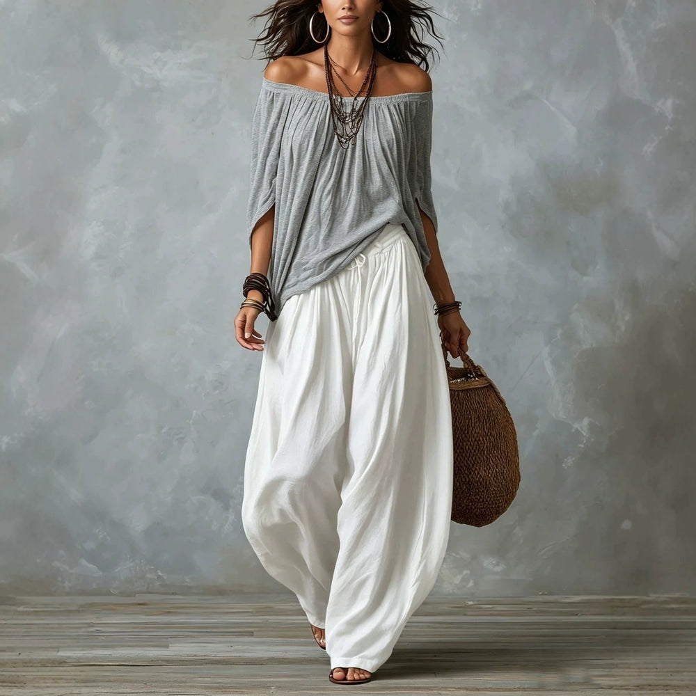 Woman wearing a gray off-shoulder top and white wide-leg pants, holding a woven bag against a gray background.