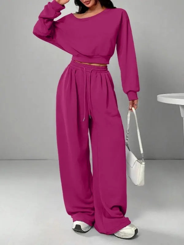 Person wearing a magenta outfit with wide-leg pants and a matching top, holding a white handbag on a gray background.