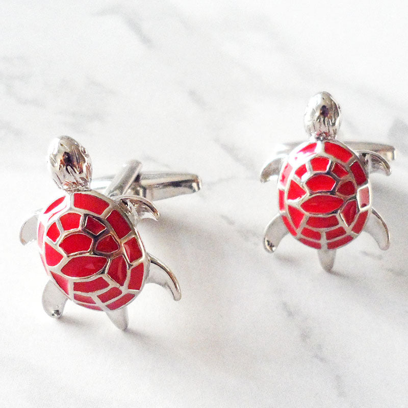 A pair of silver cufflinks shaped like small sea turtles, featuring bright red enamel scales on their shells, displayed on a light gray marble surface.