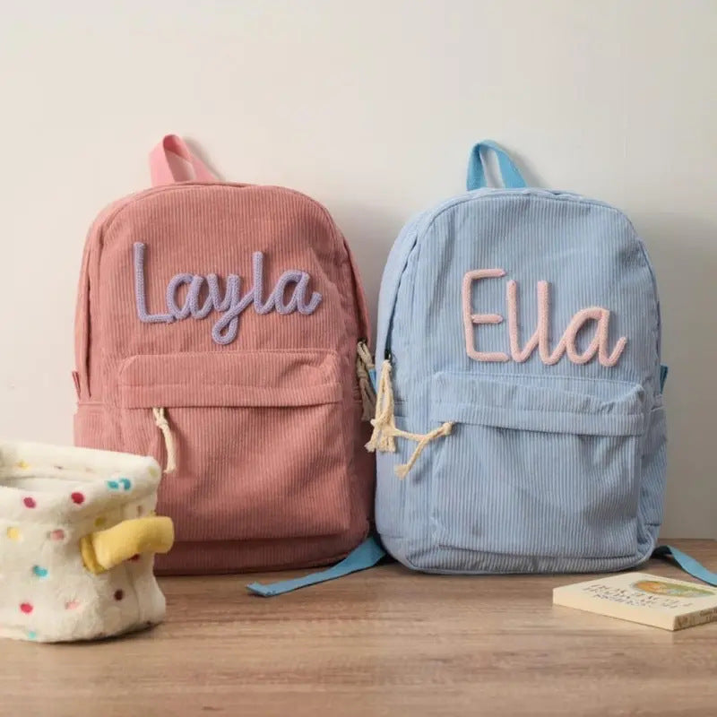 Two personalized corduroy backpacks, one dusty pink with a lavender embroidered 'Layla' and one light blue with a peach embroidered 'Ella', displayed on a wooden surface next to a polka-dot fabric basket.