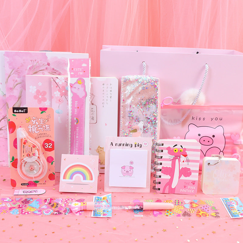A pink-themed stationery gift set featuring notebooks, a glittery phone case, a correction tape dispenser, a small notebook, a sticker pad, and a pig-themed keychain, arranged on a pink surface with a pink tulle backdrop.