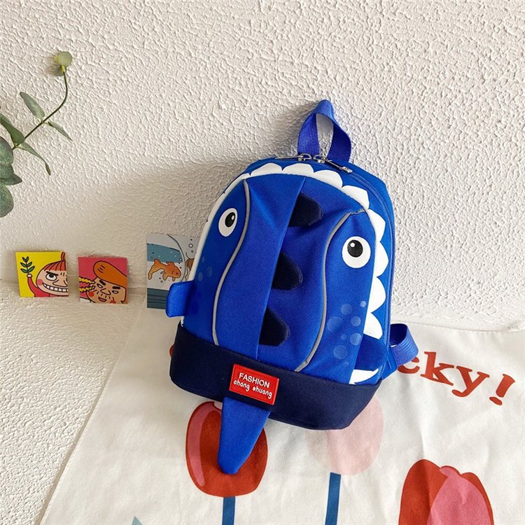 Blue shark-shaped backpack on a white wall with children's books and a plant in the background.