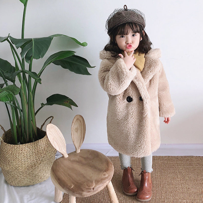 Child wearing a beige coat and hat standing next to a plant and wooden chair with rabbit ears.