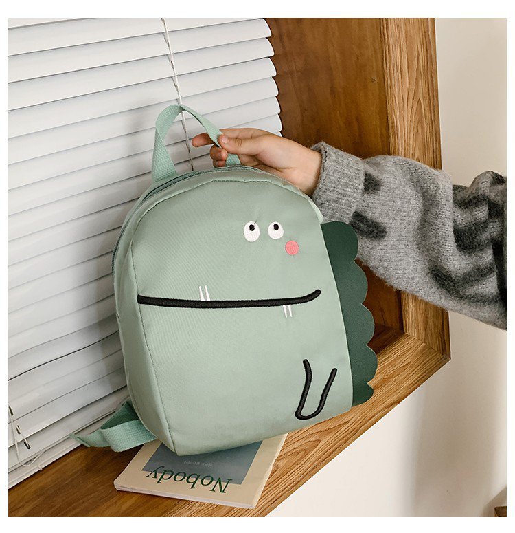 Green dinosaur-shaped backpack held by a hand on a wooden surface.