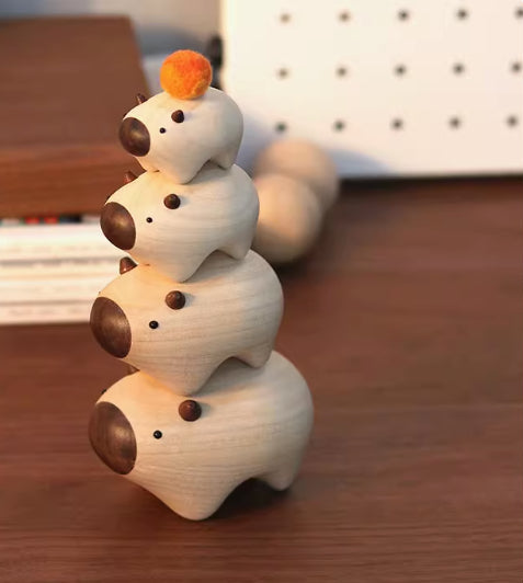 A stack of four small wooden bear figurines with dark brown snouts and noses, arranged on a wooden tabletop. The top bear has a small orange pom-pom on its head.