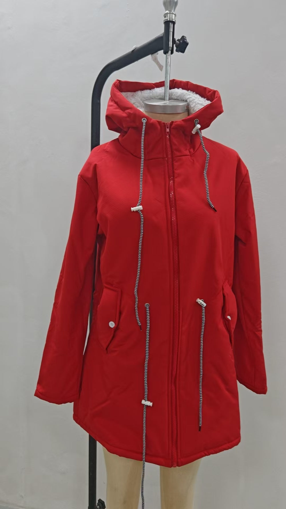 A bright red hooded parka jacket displayed on a dress form mannequin against a plain light gray wall. The jacket features a full front zipper, drawstring hood with white cord toggles, and zippered hand pockets.