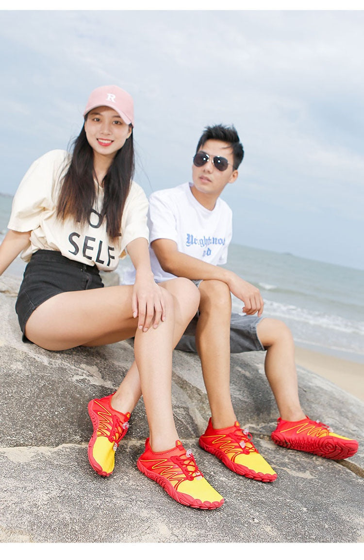 A man and a woman sitting on a large rock by the ocean, wearing bright red and yellow athletic shoes. The woman wears a cream graphic t-shirt, black shorts, and a pink cap, while the man wears a white t-shirt, sunglasses, and gray shorts.