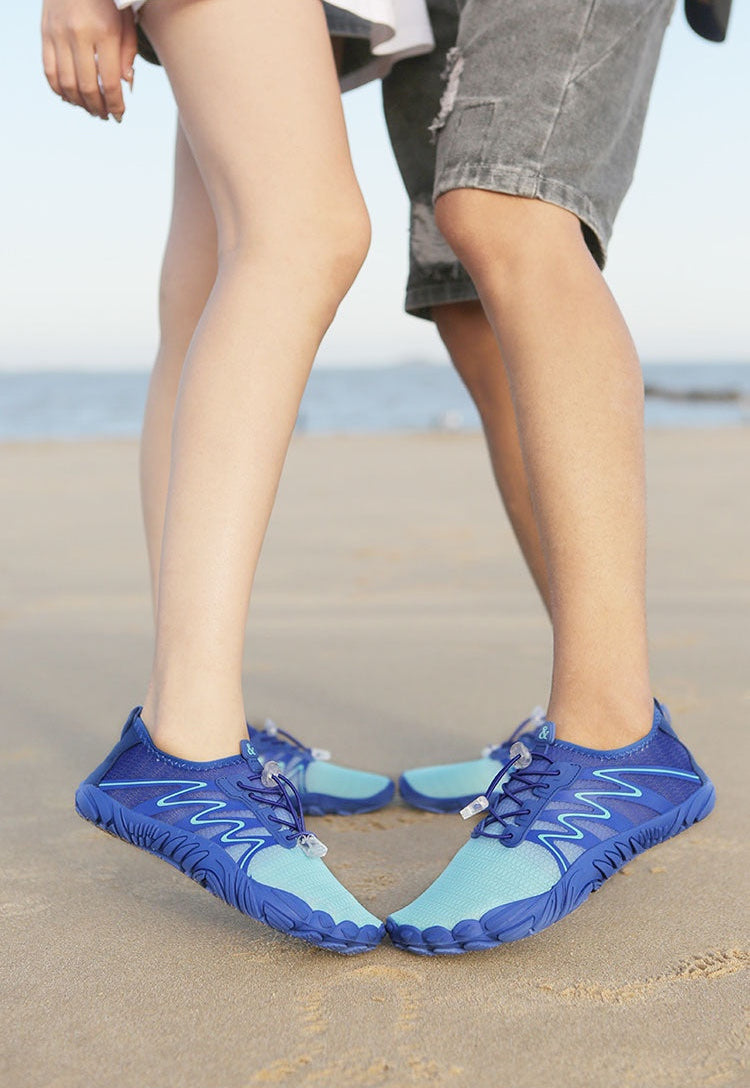 A close-up of two people standing on a sandy beach wearing blue and light blue minimalist water shoes.