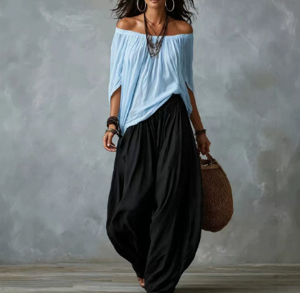 A woman wearing a light blue off-the-shoulder blouse and loose black harem pants, carrying a brown woven bag against a textured gray studio backdrop.