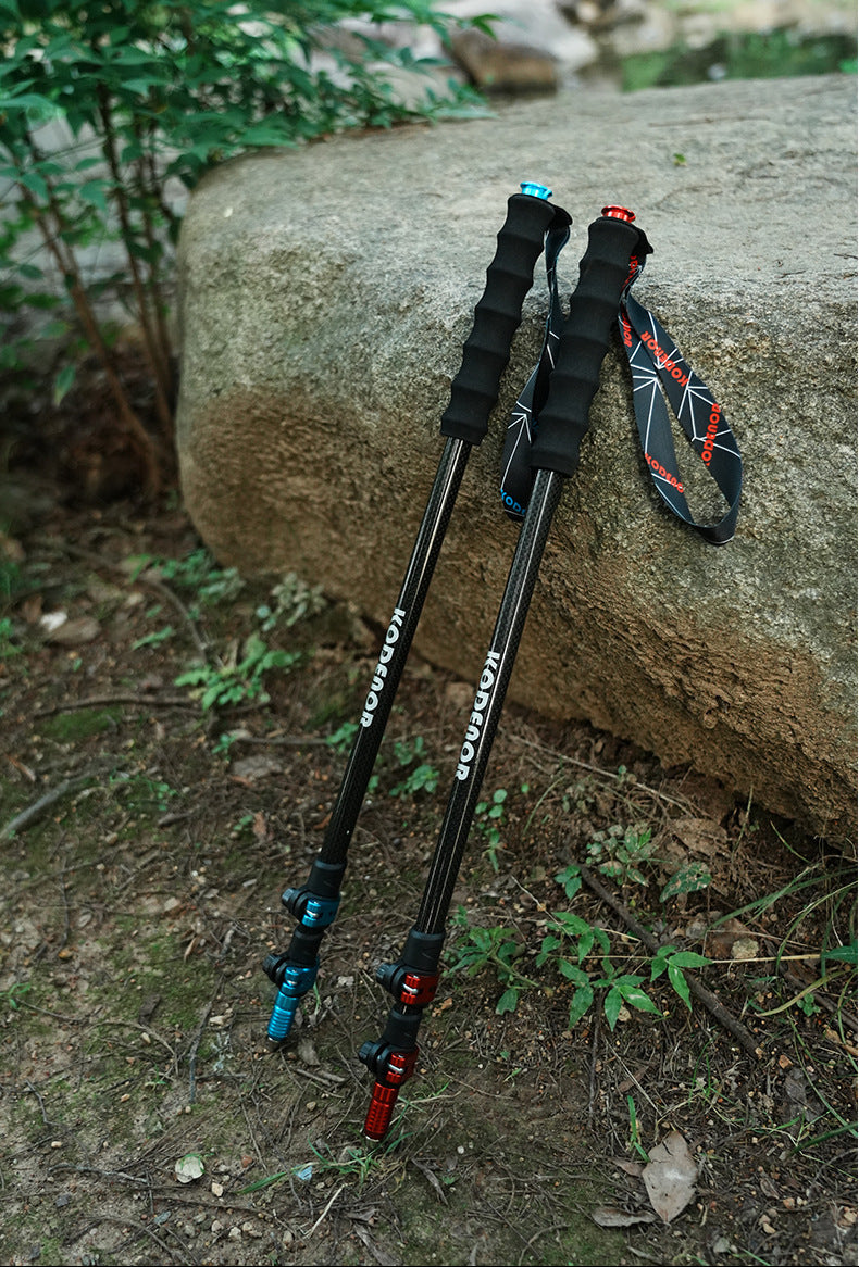 Two black hiking poles with blue and red accents, standing on a forest floor next to a large rock. A matching black and red wrist strap is draped over the rock.