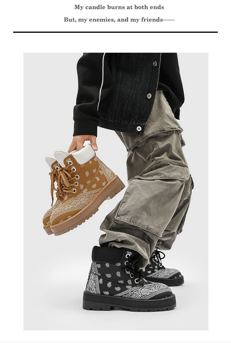 A person wearing black and gray bandana-patterned lace-up boots with gray cargo pants and a black jacket, holding a pair of tan bandana-patterned lace-up boots against a light gray background.