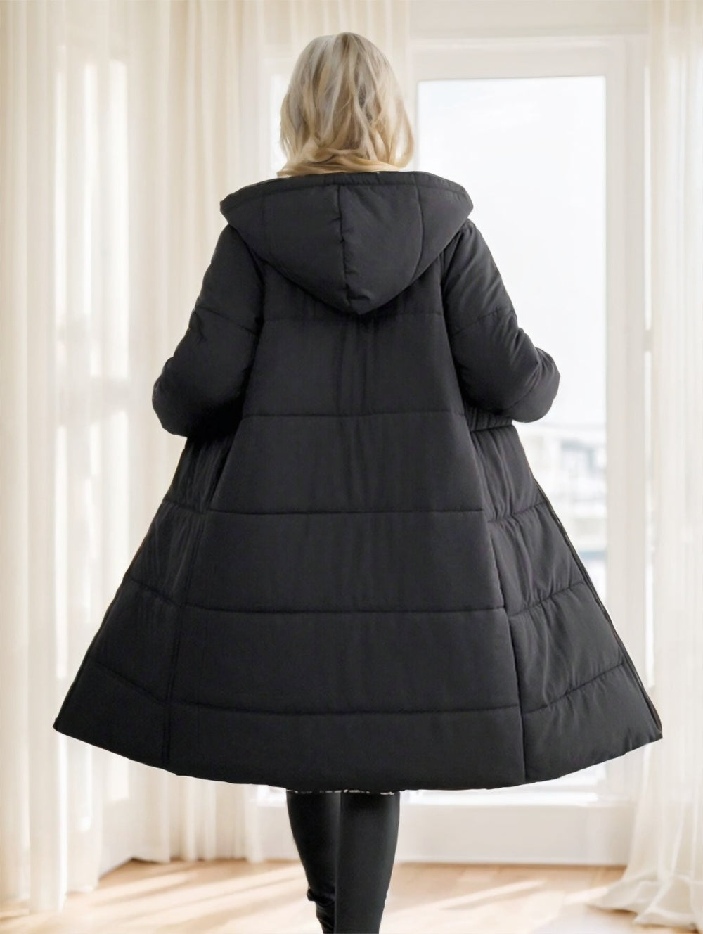 Person wearing a black puffer coat standing in a room with white curtains.