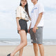 A man and a woman standing on a sandy beach wearing matching white graphic t-shirts, gray shorts, and black-and-white athletic sneakers.