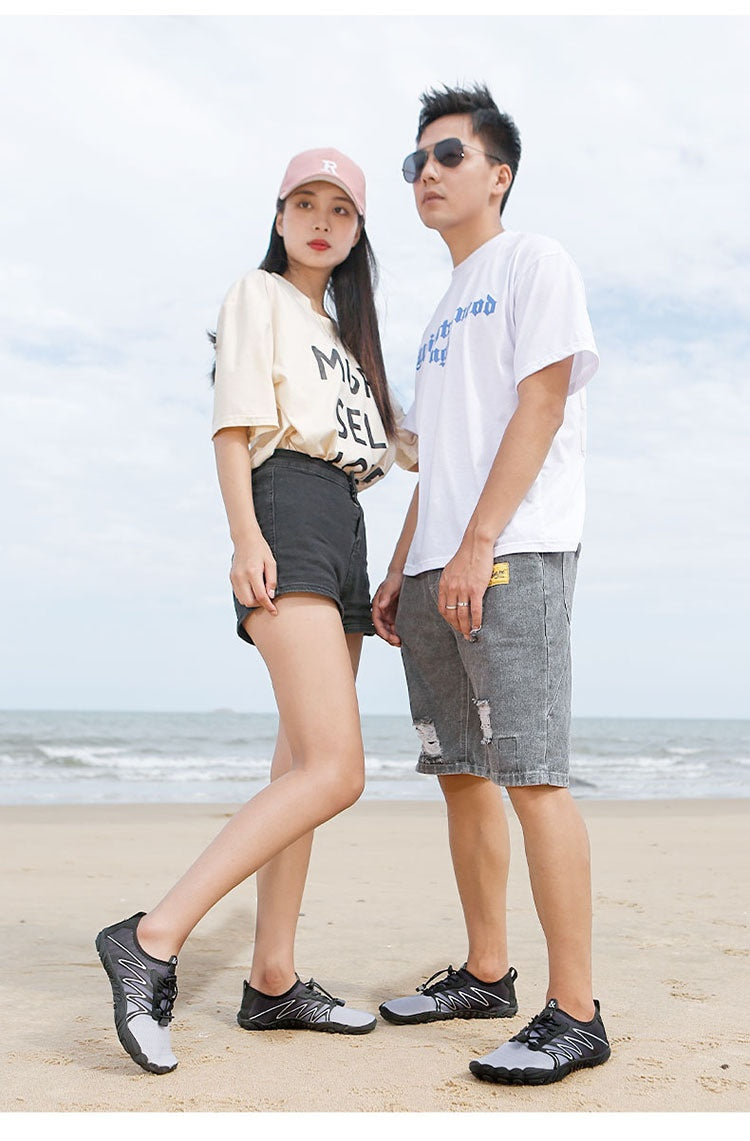 A man and a woman standing on a sandy beach wearing matching white graphic t-shirts, gray shorts, and black-and-white athletic sneakers.