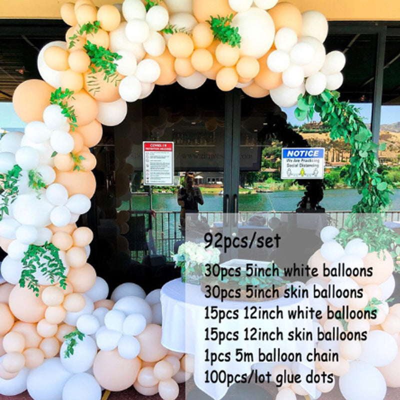 A large balloon arch decoration kit displayed in front of a glass building entrance. The arch is made of clusters of white, peach, and beige balloons with green leafy garland accents.