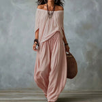A woman models a dusty pink two-piece bohemian outfit consisting of an off-the-shoulder long-sleeve top and matching loose wide-leg pants, styled with layered necklaces, bracelets, and a woven straw bag against a textured gray studio backdrop.