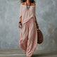 A woman models a dusty pink two-piece bohemian outfit consisting of an off-the-shoulder long-sleeve top and matching loose wide-leg pants, styled with layered necklaces, bracelets, and a woven straw bag against a textured gray studio backdrop.
