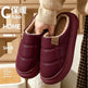 A pair of women's slip-on winter slippers in deep burgundy faux leather with a soft beige fleece lining, held by a hand wearing a beige knit sweater. The background shows a warm, cozy room interior with wooden furniture, and there are text overlays on the left side.