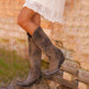 Brown knee-high boots worn with a white lace dress against a rustic stone wall.