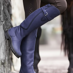 A close-up of a person wearing tall navy blue riding boots with brown leggings, standing outdoors next to a stone wall and horse legs in the background.