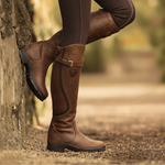 A person wearing tall brown leather riding boots with side buckles and dark leggings, seated outdoors against a stone wall with a blurred natural background.