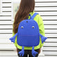 Child wearing a blue backpack shaped like a cartoon character against a white wall.