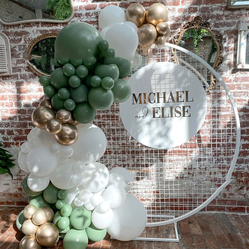Balloon arch with green, white, and gold balloons against a brick wall with 'Michael & Elise' sign.