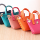 Five miniature tote handbags in navy blue, teal, pink, orange, and magenta, arranged in a diagonal row on a light wood surface.