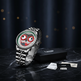 Une montre-bracelet en métal couleur argent avec un cadran rond présentant un visage de dessin animé stylisé en rouge, vert, noir et blanc, affichée verticalement à côté d'une boîte noire avec logo et de petites cartes sur une surface en marbre sombre contre un fond bleu foncé.