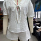 A white short-sleeve linen-like top and matching shorts set displayed on a mannequin torso in an indoor room. The top has a V-neck with a front tie detail, and the mannequin is also wearing a red cord necklace with an S pendant.