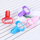 Four plastic clip-style accessories in red, purple, blue, and pink, arranged on a white textured surface with thin gray stripes and a small printed card.