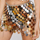 A close-up of a model wearing a short, highly reflective mini skirt covered in large metallic disc embellishments in gold, bronze, silver, and copper tones, with a chain waistband and dangling chains.