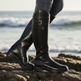 A person wearing tall black leather riding boots with a buckle strap and a small shield logo, standing on rocks with the ocean waves blurred in the background.