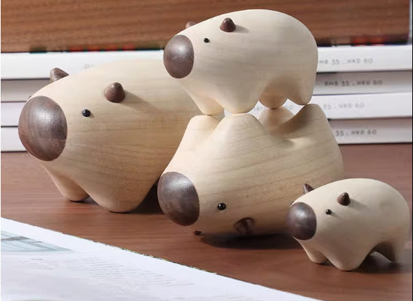 Three small wooden animal figurines with smooth natural wood bodies and dark brown snouts and ears, arranged on a wooden surface with stacked books in the background.