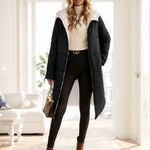 A woman models a long black puffer coat with a white sherpa collar, styled with a cream turtleneck, black skinny pants, and brown ankle boots in a bright, modern room.