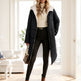 A woman models a long black puffer coat with a white sherpa collar, styled with a cream turtleneck, black skinny pants, and brown ankle boots in a bright, modern room.
