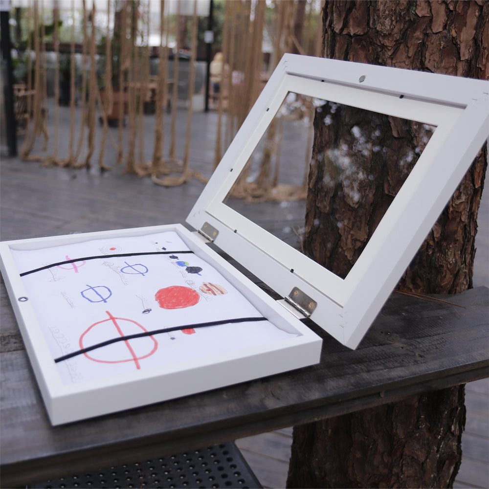 White framed picture with child's drawing leaning against a tree outdoors