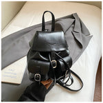 Black leather backpack held by a hand on a light-colored surface with a gray fabric background.