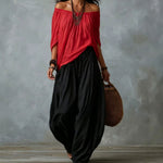 A woman wearing a red off-the-shoulder top and loose black wide-leg pants, holding a brown woven bag against a textured gray wall.