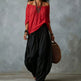 A woman wearing a red off-the-shoulder top and loose black wide-leg pants, holding a brown woven bag against a textured gray wall.