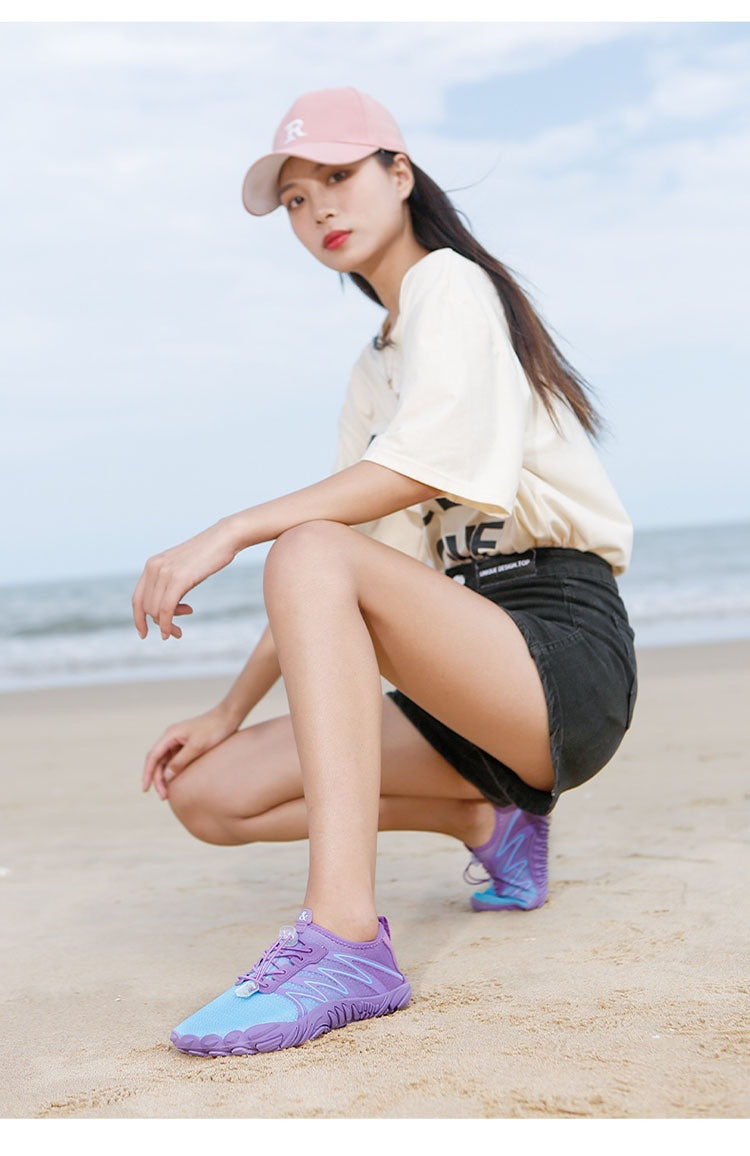 A woman crouching on a sandy beach wearing a pink baseball cap, a cream graphic t-shirt, black shorts, and purple and blue athletic shoes.