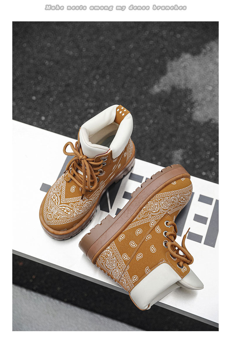 A pair of brown lace-up ankle boots with a tan bandana print overlay and white lining, displayed on a white geometric platform against a dark textured background.
